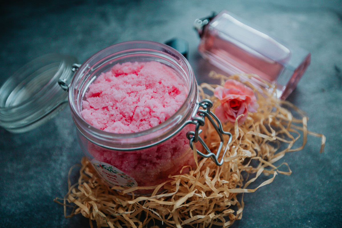 mason jar of a pink body scrub sitting on a dark gray marble counter surrounded by fake tan grass.