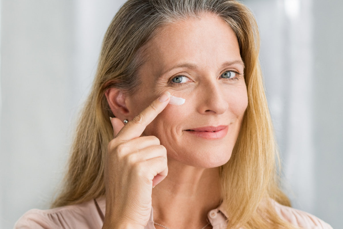 50 year old woman smiling as she applied moisturizer underneath her eye.