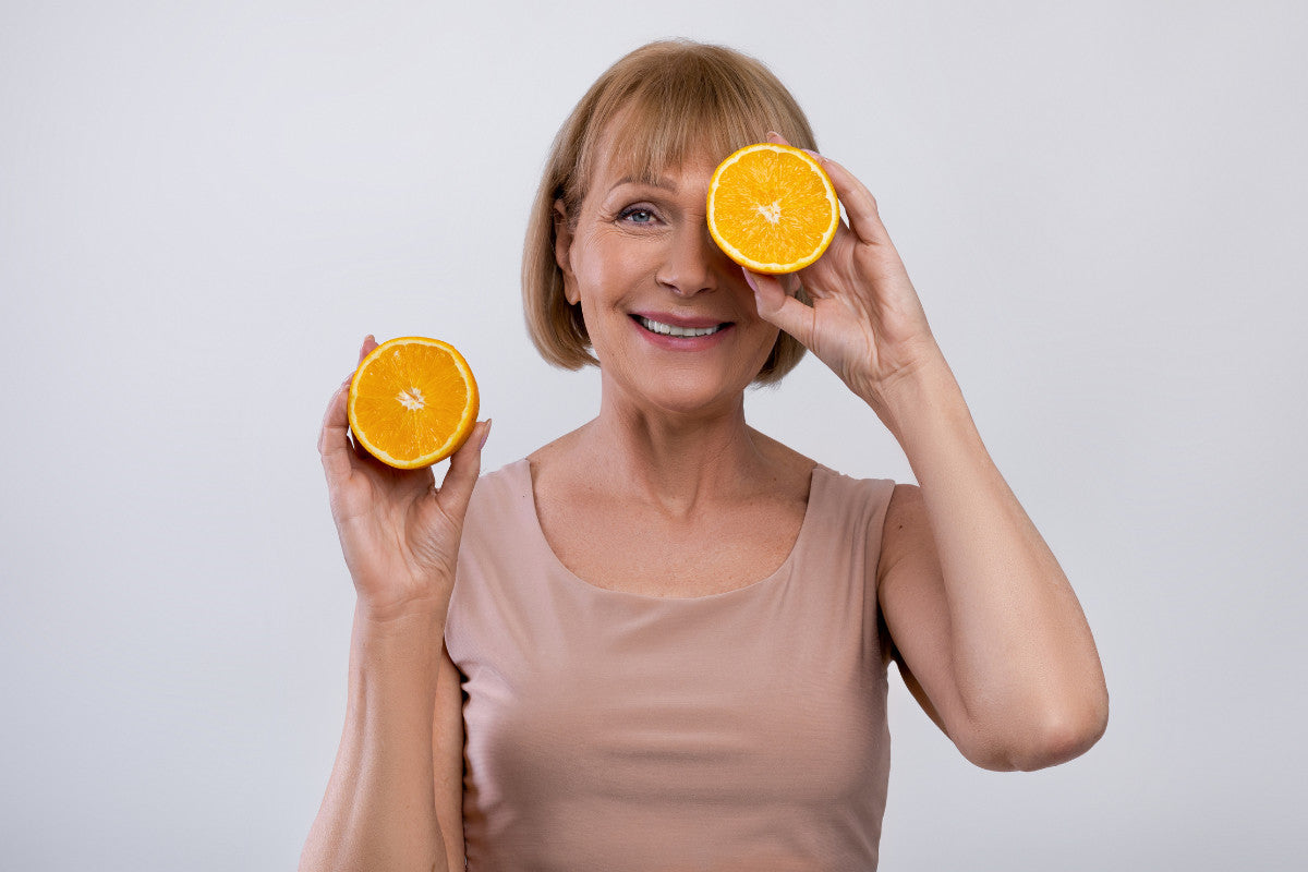 60-year-old woman holding up two halves of an orange - one is in front of her eye and the other is in her right hand.