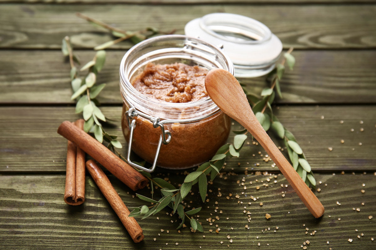 a mason jar filled with an orange-brown sugar scrub sitting on a wooden table surrounded by cinnamon sticks, a wooden spoon, and some green leaves