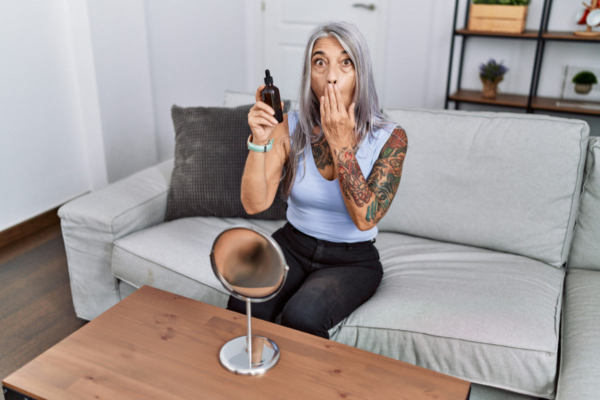 Middle age grey-haired woman using night serum sitting on the sofa covering mouth with hand, shocked and afraid for mistake.