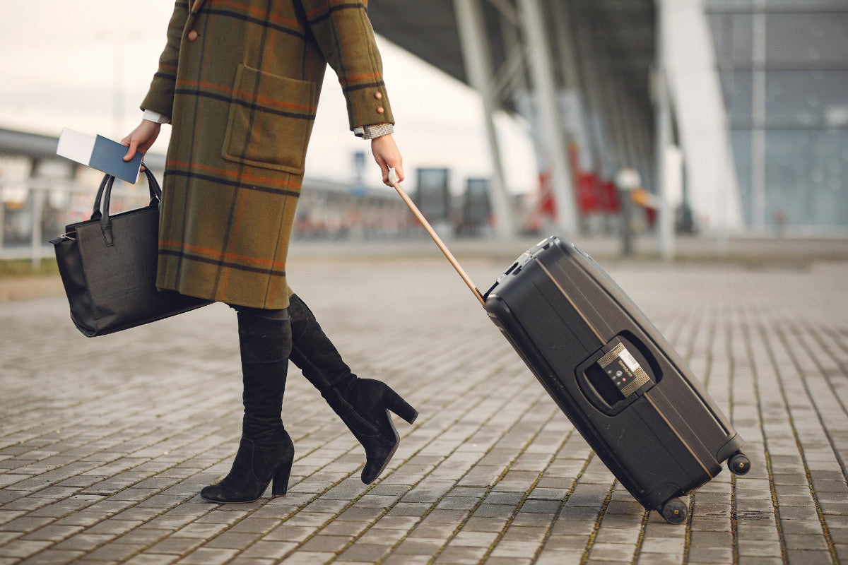 picture of the lower half of a woman wearing a plaid trench coat, high black boots, and walking outside of an airport with a black suitcase in one hand and a black purse and her passport in the other.