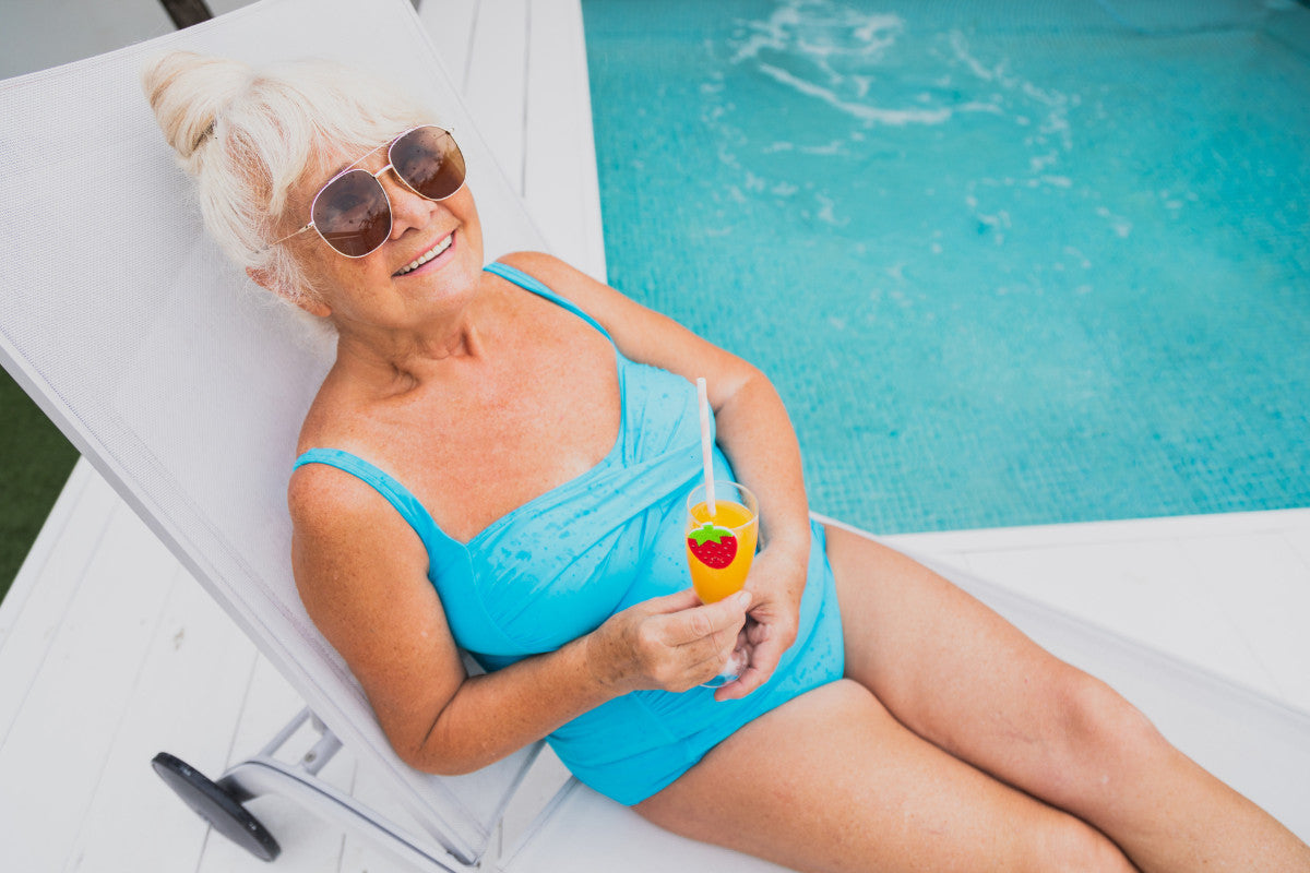 senior woman in a blue bathing suit lounging in a beach chair with a cold drink next to a pool.