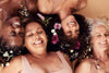 4 beautiful women of all ages and ethnicities smiling and laying next to each other with flowers in their hair