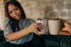 woman holding mug of hot chocolate while cheering someone else holding mug of hot chocolate