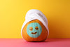 pink floor, yellow wall. orange pumpkin wearing a white towel hat, a drawn-on face in sharpie, with eyes closed and eyelashes, and a teal-green 