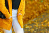 close-up of mid-section of woman wearing white pants, black shirt, and a bright orange cardigan, standing in front of autumn leaves on a pavement