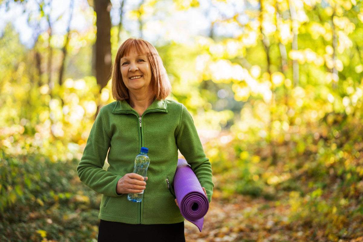 older woman smiling, wearing a green zip-up jacket, black yoga pants, and carrying a water bottle and purple yoga mat. She stands on a trail in the woods, surrounded by falling leaves during autumn.