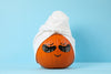 orange pumpkin with drawn on closed eyes and a smile, wearing a white towel hat and black under-eye masks, blue background