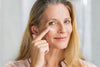 50 year old woman smiling as she applied moisturizer underneath her eye.