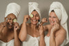 three mature woman wearing towels and towel hats, smiling and laughing as they put cucumbers over their eyes