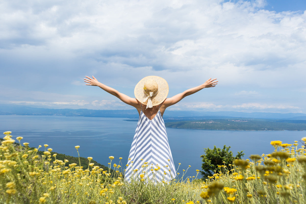 Woman in a blue and white sleeveless dress and sun hat, standing in a field of yellow wildflowers, facing away from the camera.