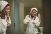 mature woman wearing a towel on her head and a white bath robe, looking at her skin in the mirror as she touches her hands to her face.