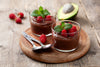 two short glasses filled with avocado chocolate mousse with raspberries on a circular wooden board.