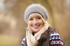 middle-aged woman smiling, bundled up in layers and sporting a gray beanie and beige scarf, sitting outside in the winter.